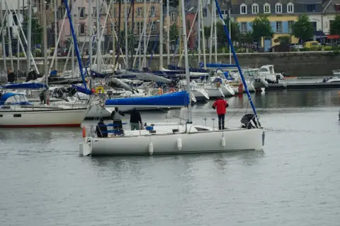 Załoga na jachcie przygotowuje się do manewru, jeden z marynarzy stoi przy sterze, inny pochyla się nad pokładem. W tle port z wieloma żaglówkami i budynkami. Szpring rufowy jest niewidoczny.