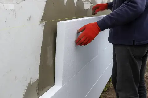 Pracownik w czerwonych rękawicach montuje płyty styropianowe do ściany. Sprawdź ocieplenie elewacji cena.
