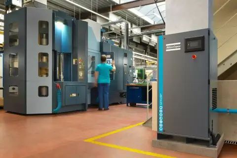 En una fábrica, un operario interactúa con una máquina industrial. A la derecha, un compresor Atlas Copco, que puede ser con aceite o sin aceite, espera su turno.