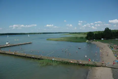 Wypływa z jeziora roś, a plażowicze kąpią się i opalają. W oddali widać żaglówki i łodzie.