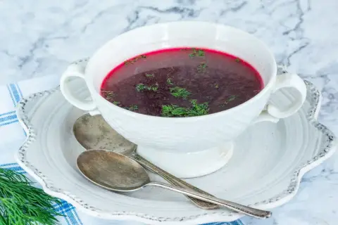Aromatic barszcz czerwony domowy, ozdobiony świeżym koperkiem, czeka w białej miseczce na talerzyku z dwoma łyżkami.