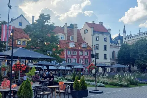 Tętniący życiem plac w Rydze, z kolorowymi kamienicami i kawiarnianymi ogródkami. W tle widać fragmenty zabytkowej architektury, przypominającej o historii państwa z Kurlandią.
