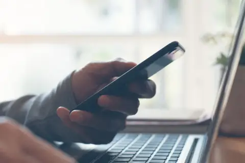 Dłoń trzyma telefon, który nie widzi laptop. Klawiatura komputera widoczna w zbliżeniu.