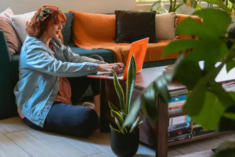 Kobieta z rudymi włosami pracuje na laptopie, siedząc na podłodze obok kanapy. To idealny przykład elastycznej **pracy hybrydowej** w domowym zaciszu.