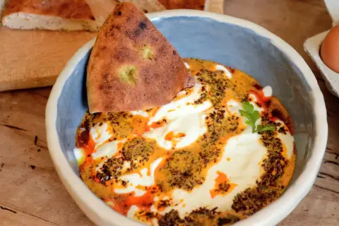 Un bol de cilbir recette, avec du pain pita trempé dans une sauce crémeuse et épicée, garni d'herbes fraîches.