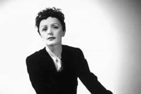 Edith Piaf w czarno-białym portrecie, w czarnym żakiecie i z charakterystyczną fryzurą.
