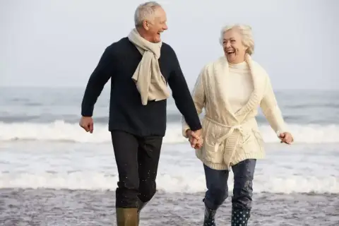 Szczęśliwa para seniorów biegnie plażą, trzymając się za ręce. Idealne tanie wycieczki dla seniorów, by poczuć radość życia!