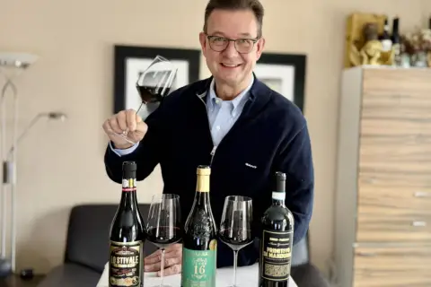 Luca Maroni Punkte: Mann mit Brille hält Weinglas, daneben drei Flaschen Rotwein und Gläser.