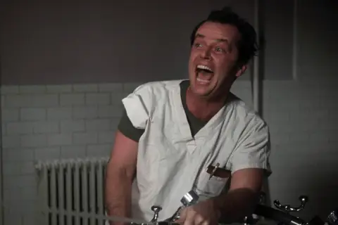 Jack Nicholson w roli McMurphy'ego w "Locie nad kukułczym gniazdem" obsada, szaleńczo krzyczy w szpitalnym ubraniu.