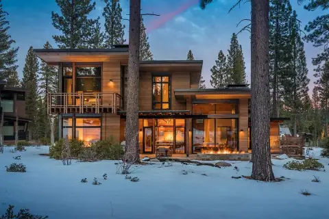 Cozy modern mountain homes with warm interior lights and a roaring fire pit, nestled amongst snow-covered trees.