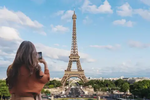 Kobieta robi zdjęcie telefonem ikonicznej wieży Eiffel. Paryski krajobraz z zielenią i budynkami w tle.