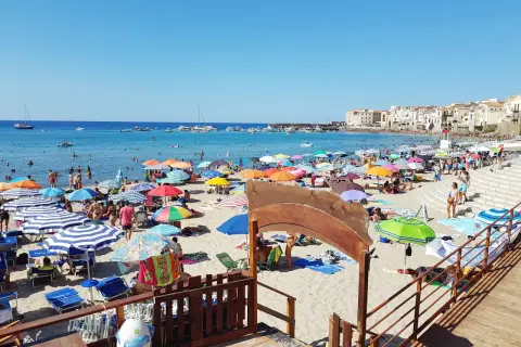 Plage animée à Cefalù en Sicile, avec des parasols colorés, des baigneurs dans la mer bleue et des bâtiments historiques en arrière-plan.