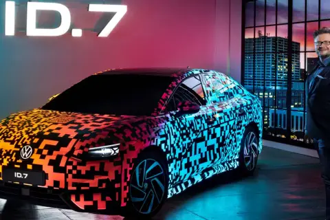 Nowy Volkswagen ID.7 prezentuje się w neonowym blasku, z efektownym wzorem QR kodu na karoserii. Obok stoi mężczyzna w garniturze, a w tle widać nocną panoramę miasta.