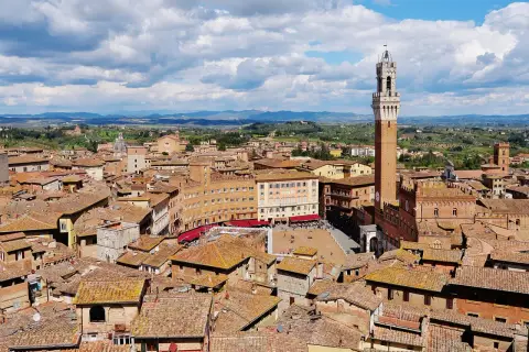Vista aerea di Siena con la Torre del Mangia che domina la Piazza del Campo, circondata da tetti rossi e colline verdi.