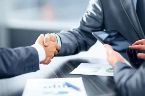 Uścisk dłoni dwóch mężczyzn w garniturach przy biurku z tabletami i wykresami.