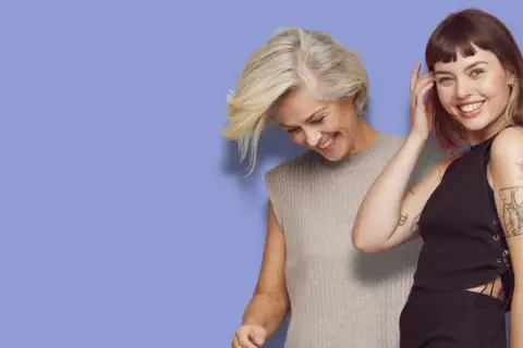 Två kvinnor med tuffa korta frisyrer ler mot kameran. Den ena har blont hår och den andra brunt.