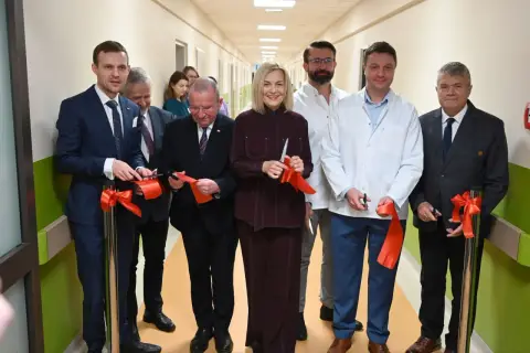 Grupa osób w garniturach i kitlach przecina czerwone wstęgi w szpitalnym korytarzu.