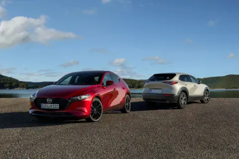 Dwie Mazdy: czerwona hatchback i beżowy SUV, stoją nad jeziorem. Sprawdź mazda cx 30 cena i wybierz swój wymarzony model.