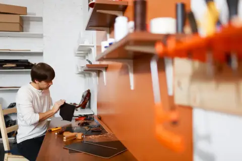 Mężczyzna pracuje w swoim atelier, tworząc skórzane wyroby. Na stole narzędzia i materiały.
