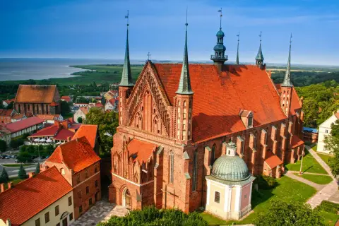 Widok na Frombork z zamkiem i katedrą. Czerwone dachy budowli kontrastują z błękitem nieba i zielenią otoczenia.