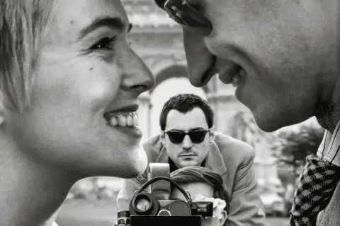 Primo piano di una donna sorridente, con un uomo che la guarda e un altro uomo con occhiali da sole che filma. Un omaggio ai registi importanti.