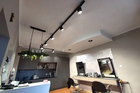 Nowoczesne oświetlenie salonu fryzjerskiego, z reflektorami na szynie i wiszącą lampą z roślinami, tworzy przytulną atmosferę.