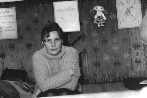 Młoda kobieta w swetrze siedzi na tle gobelinu. Na ścianie wiszą dyplomy i lalka. Czy to Agnieszka Osiecka, której prawdziwe nazwisko brzmi...