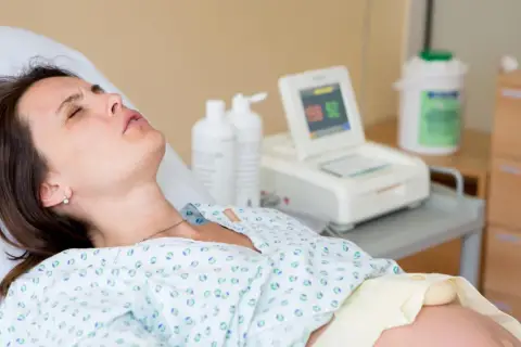 Kobieta w szpitalnym łóżku, monitor KTG, butelki z płynem. Badanie może wykluczyć obecność bakterii Streptococcus agalactiae.