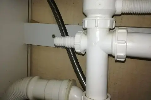 Biały plastikowy trójnik hydrauliczny pod zlewem, do którego podłączona jest elastyczna rura odpływowa i metalowy wąż.