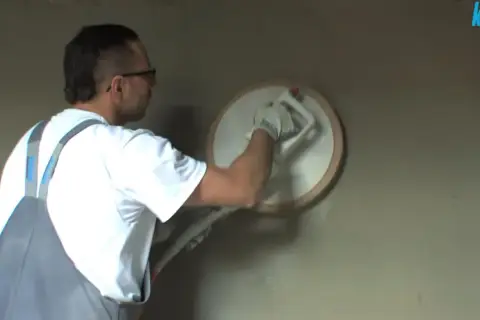 Pracownik w ogrodniczkach wykonuje zacieranie tynku cementowo wapiennego za pomocą kielni.