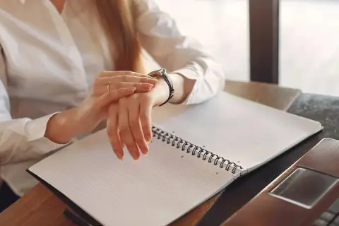 Kobieta w białej koszuli sprawdza zegarek, pracując po godzinach. Na biurku leży otwarty notatnik i laptop.