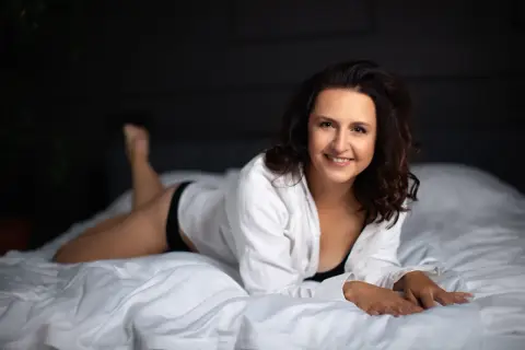 Uśmiechnięta kobieta w białej koszuli leży na łóżku. Świetne pomysły na sesję w domu.