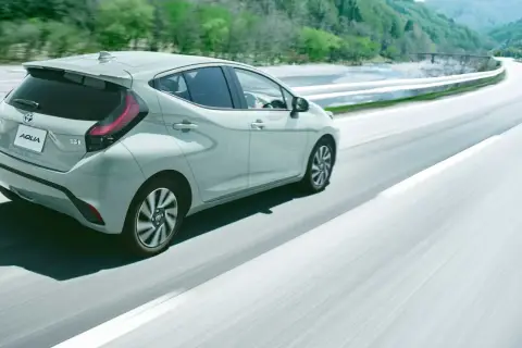 Jasnoszary Toyota Aqua jedzie po krętej drodze wśród zielonych wzgórz.