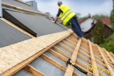 Pracownik na dachu układa blachę z membraną. Widoczny młotek i gwoździe.