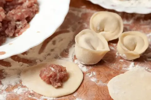 Przygotowanie pierogów z mięsem mielonym surowym. Na drewnianej desce leżą surowe pierogi, obok miska z farszem.
