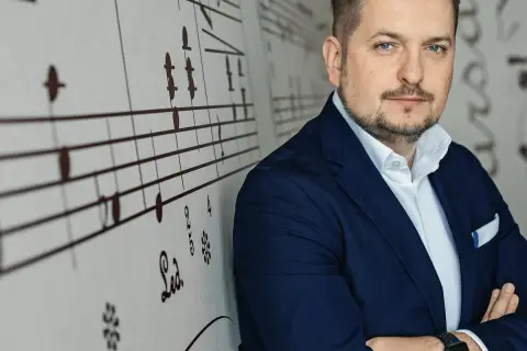Mężczyzna w granatowej marynarce, oparty o ścianę z nutami. Wygląda jak człowiek do wszystkiego, gotowy na każde wyzwanie.