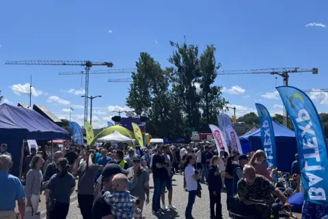 Tłum ludzi na zewnątrz, namioty, flagi i dźwigi w tle.