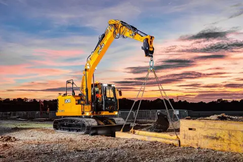 Operator koparki kurs: żółta maszyna JCB podnosi ciężki element na tle zachodzącego słońca.