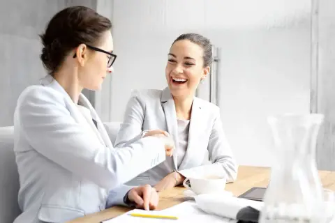 Dwie kobiety w garniturach uśmiechają się i ściskają dłonie przy biurku.
