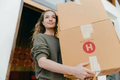 Uśmiechnięta dziewczyna z kręconymi włosami niesie dwa kartony. Na jednym z nich napis "TOYS". To idealny moment na vinted wysyłka za granicę.