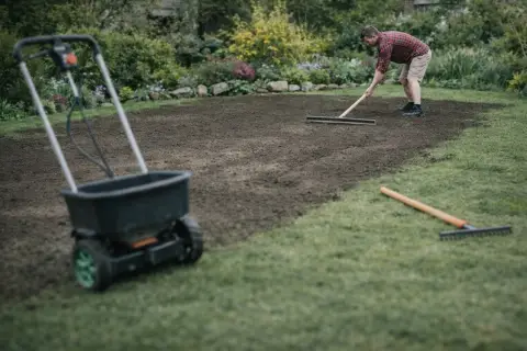 Mężczyzna zgrabia ziemię, przygotowując ją do siewu. W tle widać rozsiewacz do trawy, co sugeruje, że właśnie trwa praca nad tym, jak wyrównać trawnik.