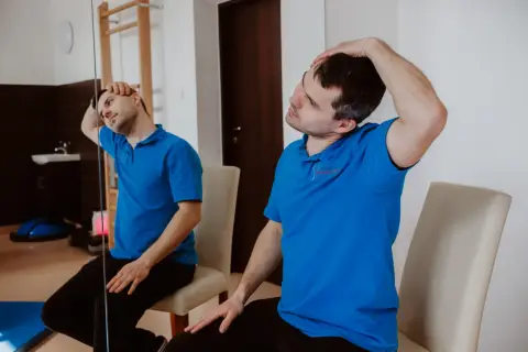 Mężczyzna w niebieskiej koszulce wykonuje ćwiczenia na kręgosłup szyjny, delikatnie przechylając głowę do boku.