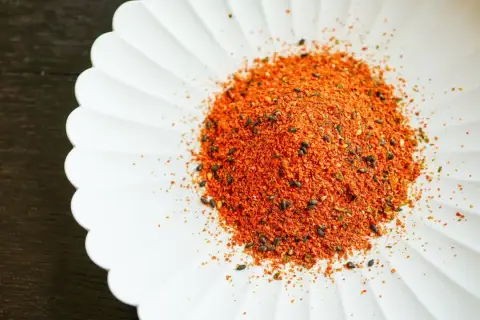 A pile of vibrant Japanese 7 spice blend, featuring chili flakes and black sesame seeds, sits on a white, fluted dish.