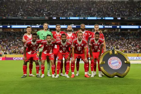 Drużyna Bayern Monachium – zawodnicy w czerwonych strojach pozują do zdjęcia na tle trybun. Obok nich stoi okrągłe logo klubu.