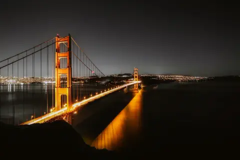 Złota Brama nocą, oświetlona, nad wodą. W oddali widać miasto w Kalifornii, San Francisco, z migoczącymi światłami.
