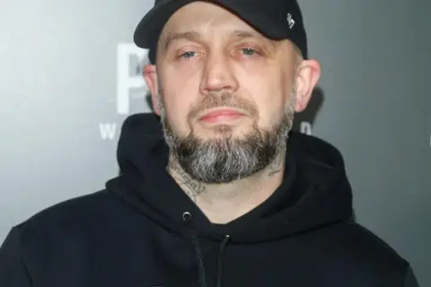 Mężczyzna w czarnej bluzie z kapturem i czapce z daszkiem, z brodą i tatuażami na szyi. Pikers ile ma lat?