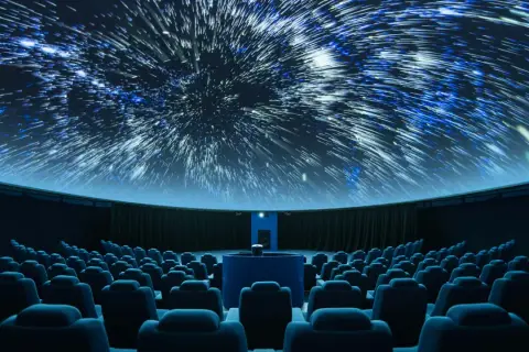 Wnętrze planetarium, gdzie na kopule wyświetlane są rozgwieżdżone niebo i galaktyki. Widownia z niebieskimi fotelami czeka na seans.