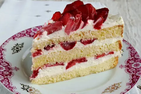 Pyszny tort z bitą śmietaną i truskawkami, przełożony kremem i owocami, na ozdobnym talerzyku.