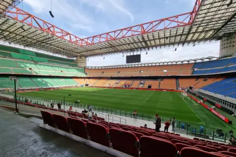 Pusta trybuna stadionu San Siro z zieloną murawą i pomarańczowymi oraz niebieskimi siedzeniami.