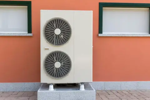 Unidad exterior de bomba de calor con dos ventiladores, instalada junto a una pared naranja con dos ventanas.